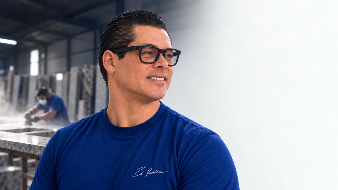 Zé Poeira, de camiseta azul, dentro de uma marmoraria com chapas de granito ao fundo.